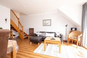 Un salon moderne avec un escalier en bois, un canapé d’angle, une table basse en bois et un fauteuil en osier.