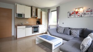 A modern kitchen with white cabinets, a microwave, and a built-in oven. There is a gray sofa with decorative pillows and a glass coffee table in the living area. - Urlauberdorf Haus 63a (Ostseebad Boltenhagen)