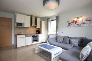 A modern kitchen with white cabinets, a microwave, and a built-in oven. There is a gray sofa with decorative pillows and a glass coffee table in the living area.