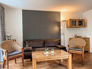 Een woonkamer met een bruine leren bank, twee rieten fauteuils, een houten salontafel en een behang met patroon.