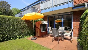 A patio with outdoor furniture and a yellow umbrella, a brick building with a blue balcony, and a well-maintained hedge. - Papillon Wohnung 04-2 (Ostseebad Boltenhagen)