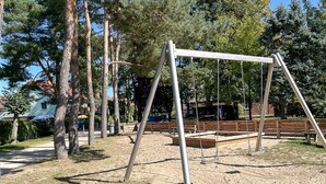 A playground with swings and benches, surrounded by trees and a building in the background. - Papillon Wohnung 04-2 (Ostseebad Boltenhagen)