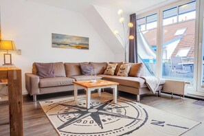 Ein modernes Wohnzimmer mit einer Couch, einem Couchtisch, einem Teppich mit Kompassmuster und großen Fenstern mit Blick ins Freie.