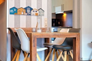 A dining area with a wooden table and chairs, a kitchen in the background, and a wall with house-shaped decorations. - Papillon Wohnung 04-6 (Ostseebad Boltenhagen)
