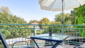 Un balcon avec une table en verre, deux chaises, une plante en pot et un parasol blanc.