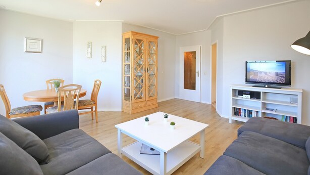 Un salon avec une table basse blanche, des canapés gris, un meuble en bois, une table à manger avec des chaises et un téléviseur posé sur un meuble blanc.