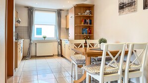 A modern kitchen with a dining area, featuring a wooden table, white chairs, and a bookshelf.