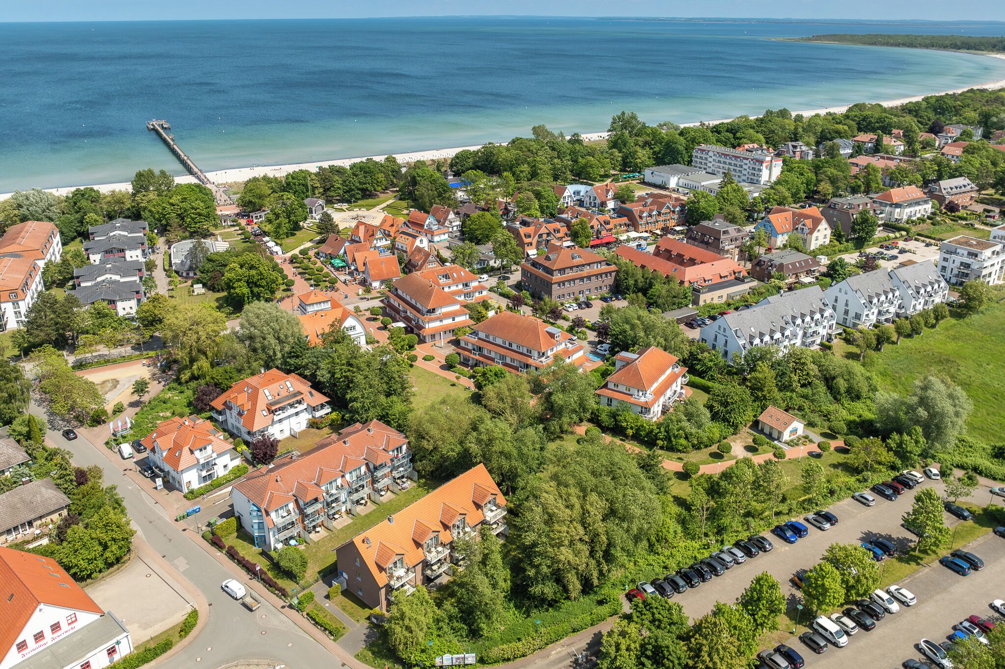 An aerial view of a coastal town with a mix of residential and commercial buildings, a pier extending into the sea, and a beach in the background.
