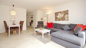 A modern living room with a grey sofa, red cushions, a white coffee table, and a dining area with a white table and brown chairs. - Papillon Wohnung 05-2 (Ostseebad Boltenhagen)