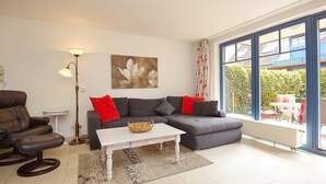 A living room with a sectional sofa, a coffee table, a floor lamp, and a large window with a view of the outdoors. - Papillon Wohnung 05-2 (Ostseebad Boltenhagen)