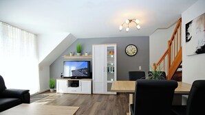 A modern living room with a TV, wooden furniture, a staircase, and a clock on the wall. - Papillon Wohnung 05-6 (Ostseebad Boltenhagen)