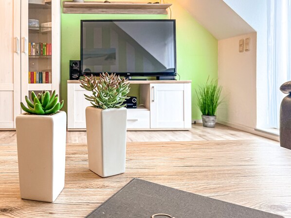 A modern living room with a flat-screen TV, two potted plants, a bookshelf, and a wooden floor.