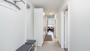 Un couloir étroit aux murs blancs, avec un banc et une porte donnant sur un espace de vie.