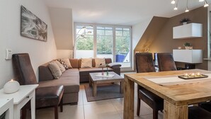 Un salon moderne avec un canapé, une table basse et une table à manger. Une fenêtre laisse entrer la lumière naturelle et offre une vue sur la verdure environnante.