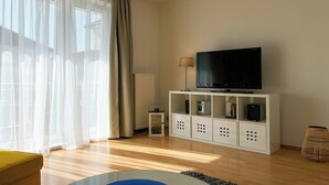 A modern living room with a flat-screen TV on a white cabinet, a yellow sofa, and a large window with sheer curtains. - Waterkant - Muschel Wohnung 1 (Ostseebad Boltenhagen)
