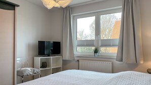 A bedroom with a bed, a TV on a stand, a chair, a window with curtains, and a radiator. - Waterkant - Strandhafer Wohnung 6 (Ostseebad Boltenhagen)