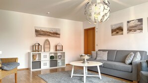 A modern living room with a grey sofa, a white coffee table, and a grey rug. There are wall-mounted shelves with decorative items and framed pictures. - Waterkant - Strandhafer Wohnung 6 (Ostseebad Boltenhagen)