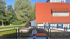 A balcony with a glass table and chair, overlooking a garden and a building with a red roof. - Waterkant - Strandhafer Wohnung 6 (Ostseebad Boltenhagen)