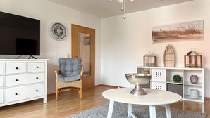 A modern living room with a white TV stand, a grey armchair, a round table with a silver bowl, and a decorative wall with a painting and birdcages. - Waterkant - Strandhafer Wohnung 6 (Ostseebad Boltenhagen)