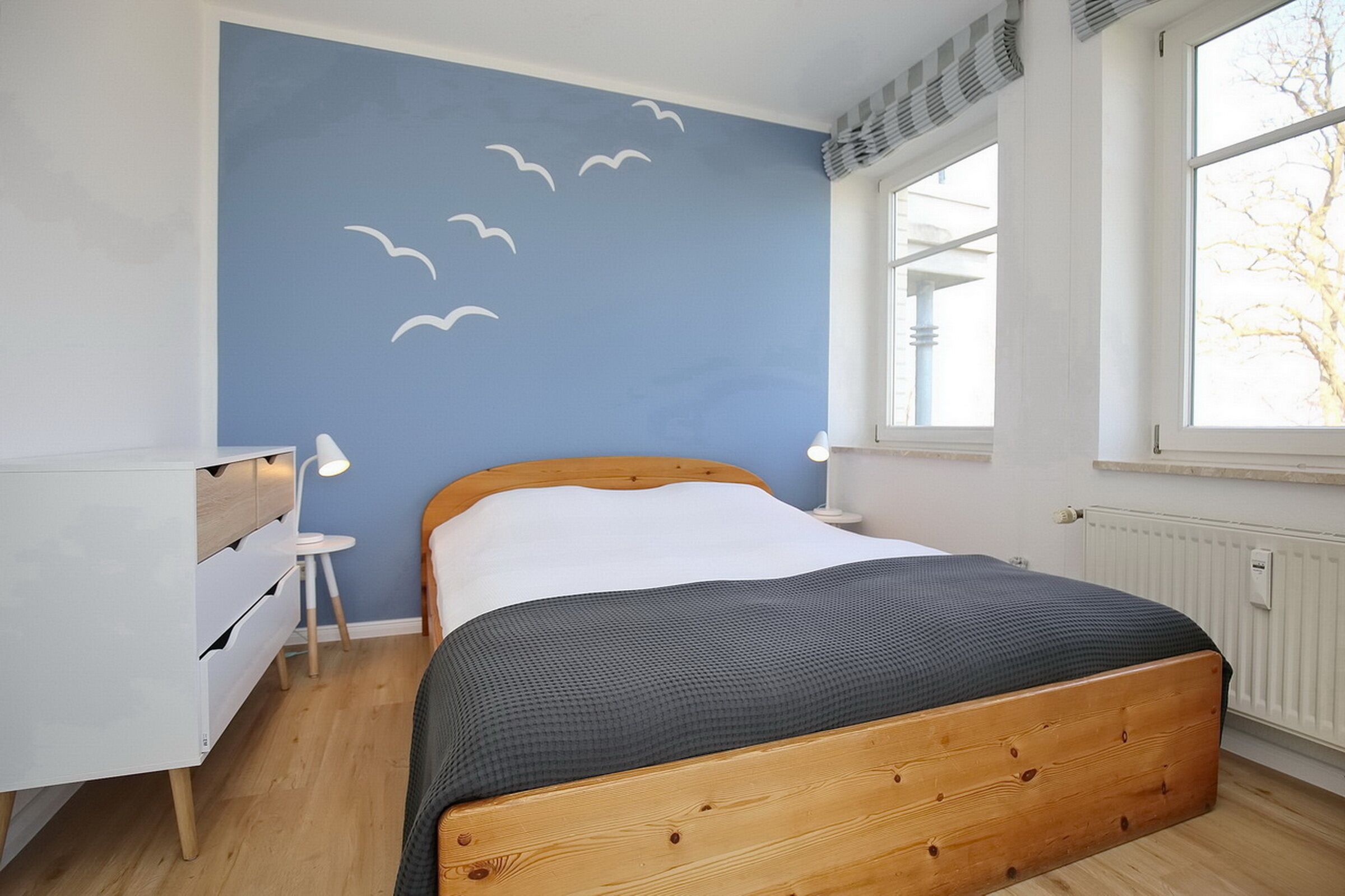 Une chambre à coucher avec un lit en bois, une commode blanche, une table de chevet et une fenêtre donnant sur des arbres.