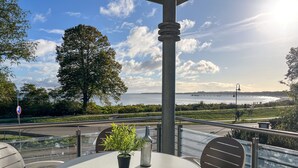 Une table ronde, entourée de chaises, et un pot de fleurs sont placés sur un balcon qui donne sur un plan d’eau et sur un pont situé au loin.