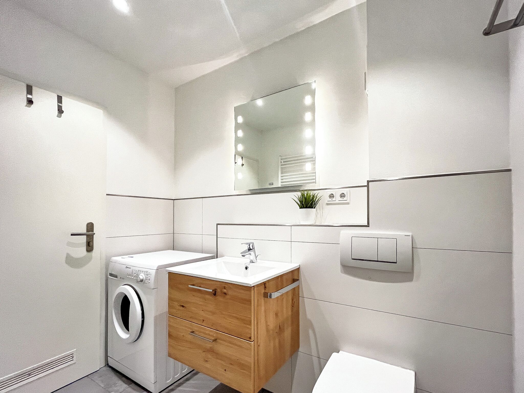 Ein modernes Badezimmer mit integrierter Waschmaschine, einem Holz-Waschtisch, einem Spiegel mit Beleuchtung und einer Toilette.
