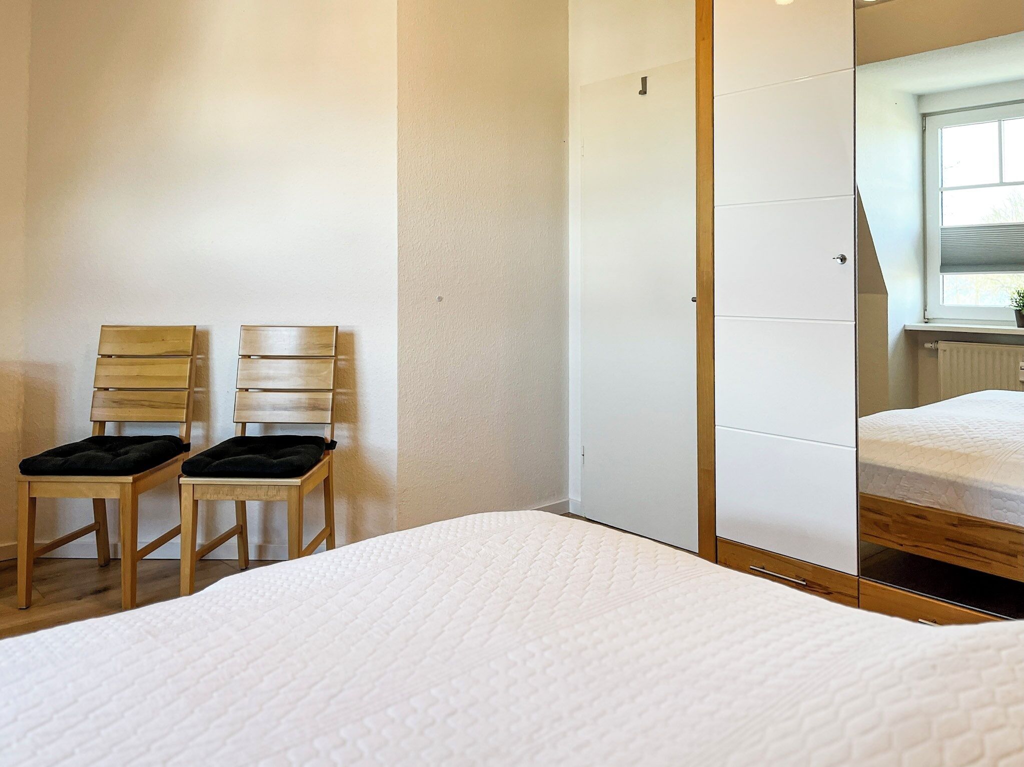 A bedroom with a bed, two wooden chairs with black cushions, a wardrobe, and a window with blinds.
