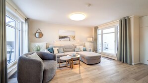 A spacious living room with a grey sectional sofa, a round coffee table, and a large window with curtains. - Haffblick Wohnung 50 (Ostseebad Boltenhagen)