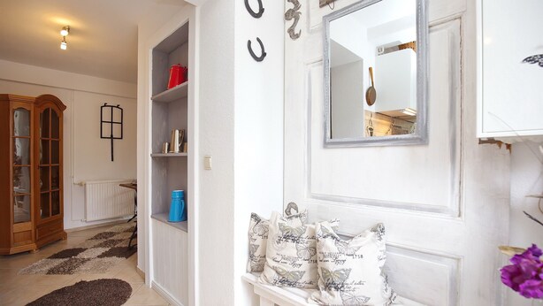 A hallway with a white door, a mirror, a blue bucket, and a patterned pillow. - Einliegerwohnung Arielle 1 (Boltenhagen/Redewisch)