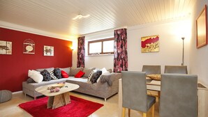 A living room with a red wall, grey sofa, and a dining area with a table and chairs. - Einliegerwohnung Arielle 1 (Boltenhagen/Redewisch)