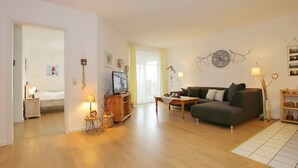 A spacious living area with a sofa, a TV, and a wooden coffee table. There is a door leading to a bedroom and a window with curtains. - Dünenblick Wohnung 05 (Ostseebad Boltenhagen)