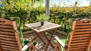 A wooden outdoor seating area with a table and chairs, surrounded by lush greenery and a clear sky. - Dünenblick Wohnung 05 (Ostseebad Boltenhagen)
