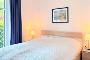 Una cama bien hecha con colcha blanca, un cuadro enmarcado en la pared y dos lámparas de noche.