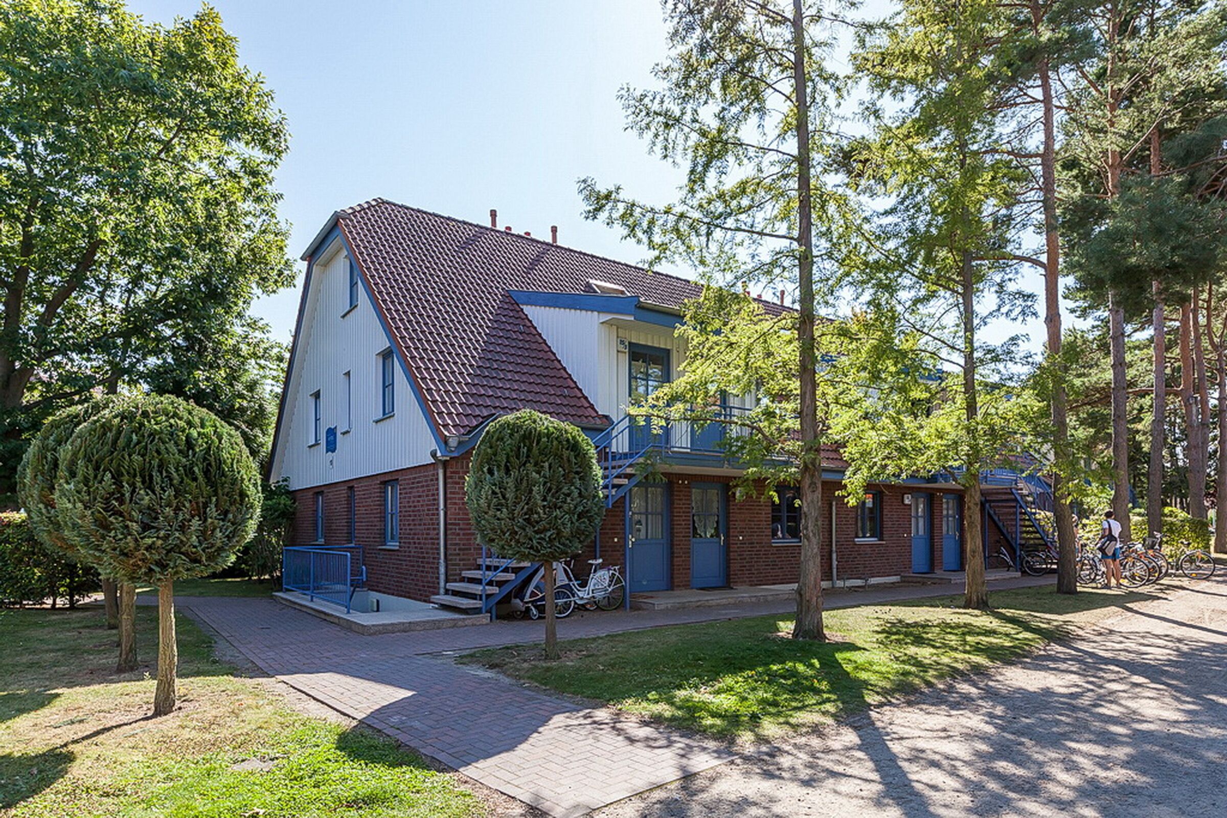 Een tweelaags huis met een rood pannendak, een blauwe voordeur en een klein portaaltje. Buiten staan bomen en staat een fiets geparkeerd.