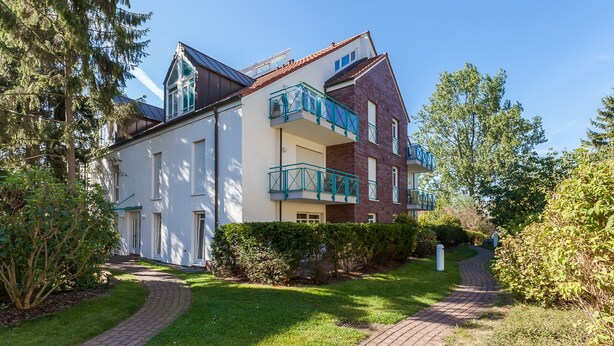 Um edifício residencial de vários andares com fachada de tijolo, varanda com grades azuis e um jardim bem cuidado com um caminho pavimentado.