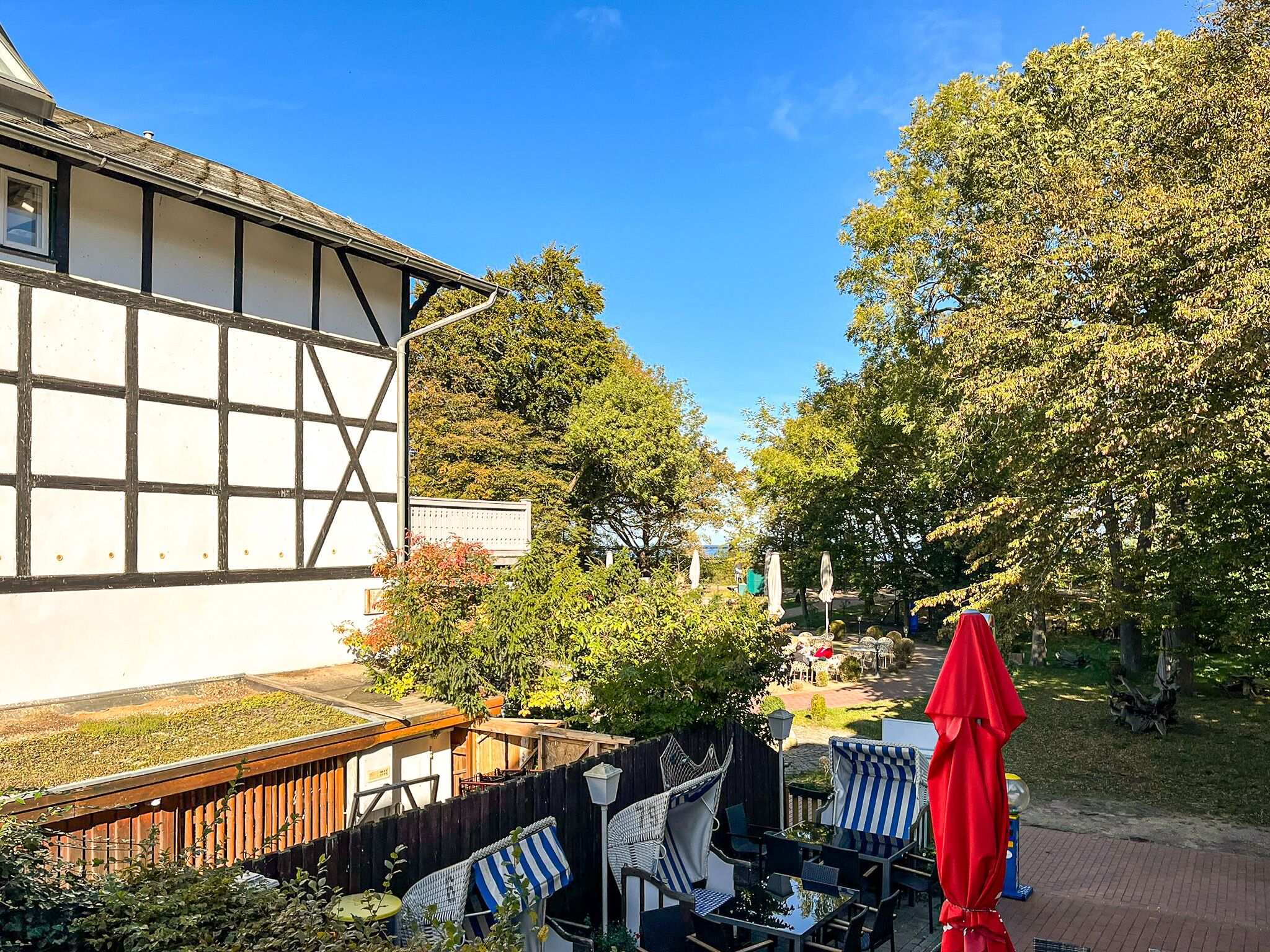 Ein traditionelles Gebäude mit heller Fassade und dunkler Umrandung, neben einem gepflegten Garten mit Außenmöbeln und Sonnenschirmen.