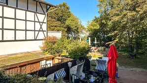 Ein traditionelles Gebäude mit heller Fassade und dunkler Umrandung, neben einem gepflegten Garten mit Außenmöbeln und Sonnenschirmen.