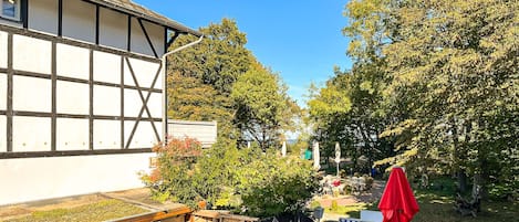 Ein traditionelles Gebäude mit heller Fassade und dunkler Umrandung, neben einem gepflegten Garten mit Außenmöbeln und Sonnenschirmen.