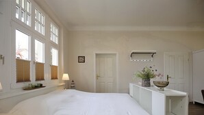 Une chambre avec un grand lit, une table de chevet, une commode, une coiffeuse, une fenêtre avec des stores, une porte et une étagère murale ornée d’un vase et d’un bol.