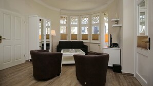 Un salon avec un canapé noir, deux fauteuils marron, une table basse blanche et une baie vitrée avec des volets blancs.