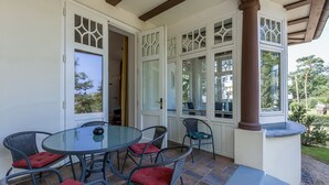 Une terrasse couverte avec une table en verre et des chaises en osier, une porte blanche ornée de panneaux de verre décoratifs, et une vue sur la verdure environnante.