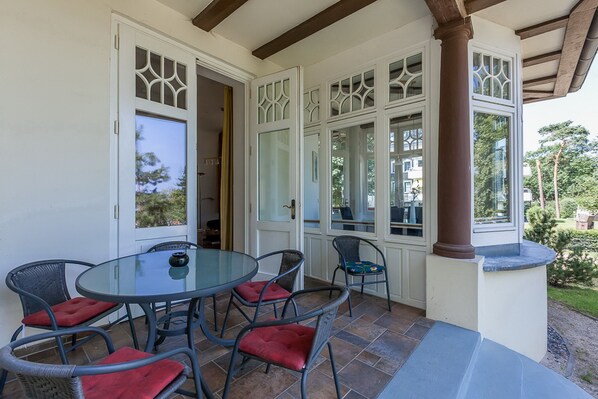 A covered patio with a glass table and wicker chairs, a white door with decorative glass panels, and a view of the surrounding greenery.