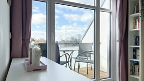 Ein Zimmer mit einem großen Fenster, einem weißen Tisch mit einem Glasbehälter, einem Bücherregal und Blick auf einen Balkon mit Stühlen.