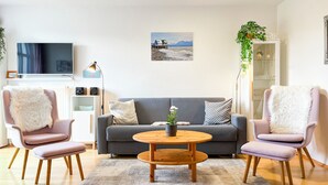 Ein modernes Wohnzimmer mit einem grauen Sofa, zwei rosa Sesseln, einem Holztisch und einem an der Wand befestigten Fernseher.