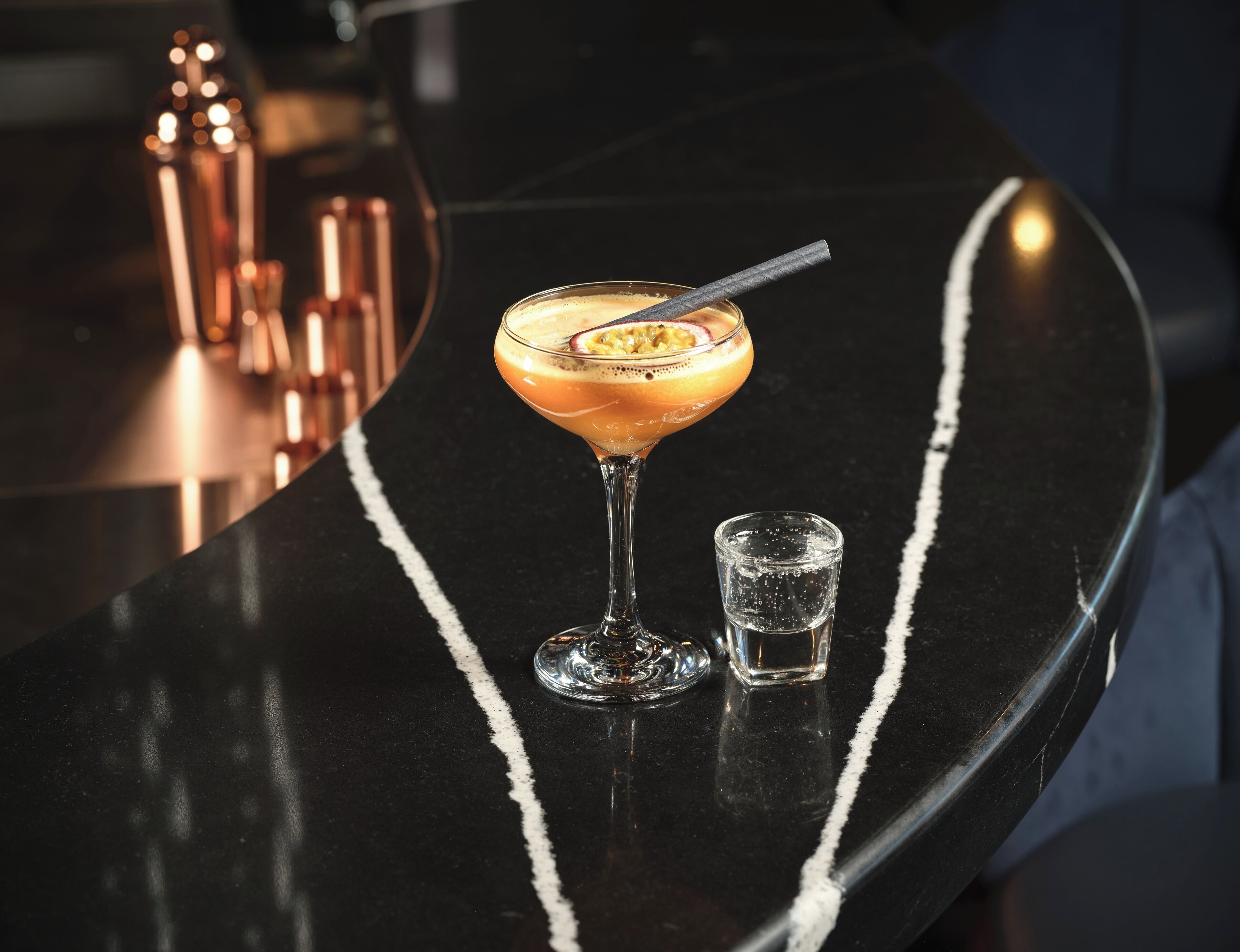 A cocktail on a black bar counter with a small glass of water beside it.