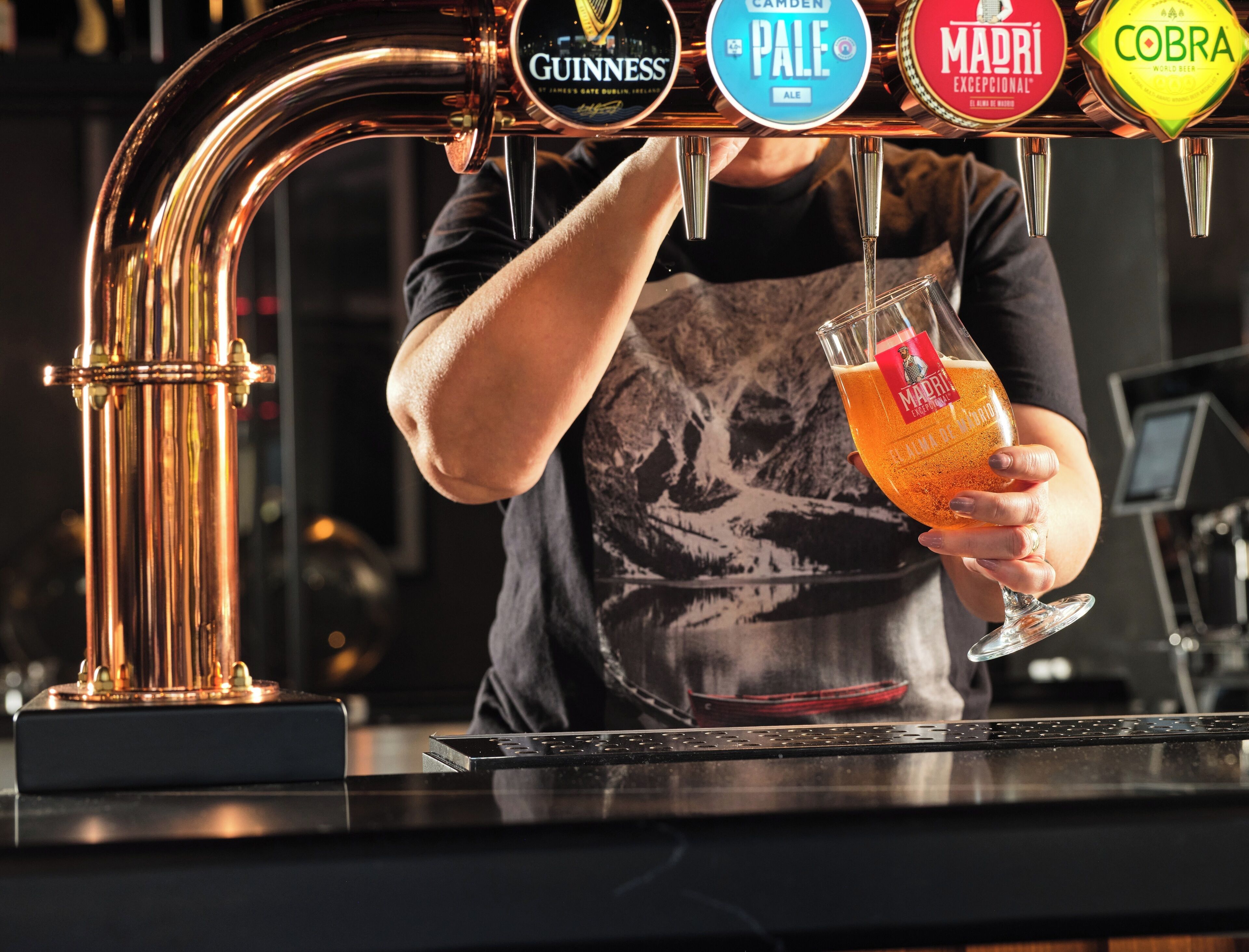 A bartender pouring a drink from a tap labeled with brands like Guinness, Pale Ale, Madri, and Cobra.