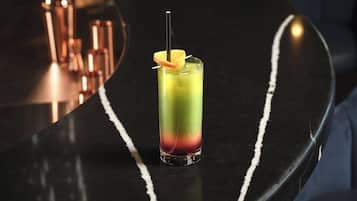 A tall glass of layered drink with a straw and an orange slice garnish on a black bar counter.