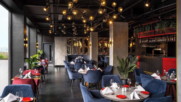 A modern restaurant interior with round tables, blue upholstered chairs, and a bar area with a red accent wall. - Radisson RED London Gatwick Airport (Crawley)