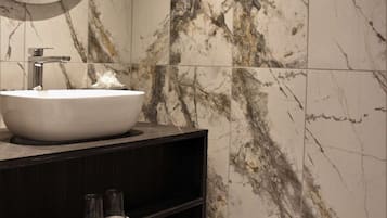 Un baño con salpicadero de mármol, un lavamanos blanco, un mueble de tocador de madera oscura y toallas enrolladas ordenadamente.