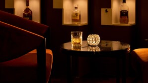 Een bar met planken waarop flessen staan, een ronde tafel met een glas en een decoratief object, en een rode stoel.